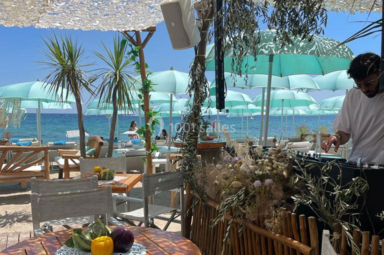 Terrasse en bord de mer avec parasols verts, tables en bois, décoration végétale et un DJ à droite.