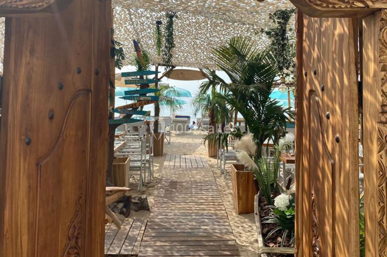 Entrée en bois sculpté menant à une plage avec des parasols, des plantes et une vue sur la mer turquoise.