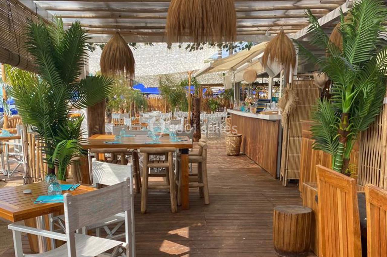 Terrasse en bois d'un restaurant avec tables, chaises, plantes tropicales et luminaires suspendus en paille.