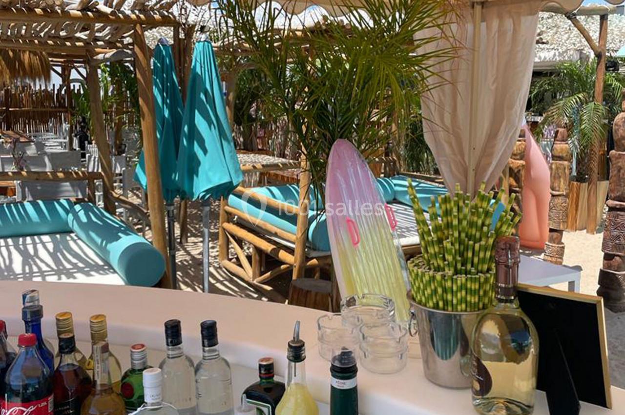 Bar extérieur avec bouteilles, verres et décor tropical, entouré de transats, parasols et végétation exotique.
