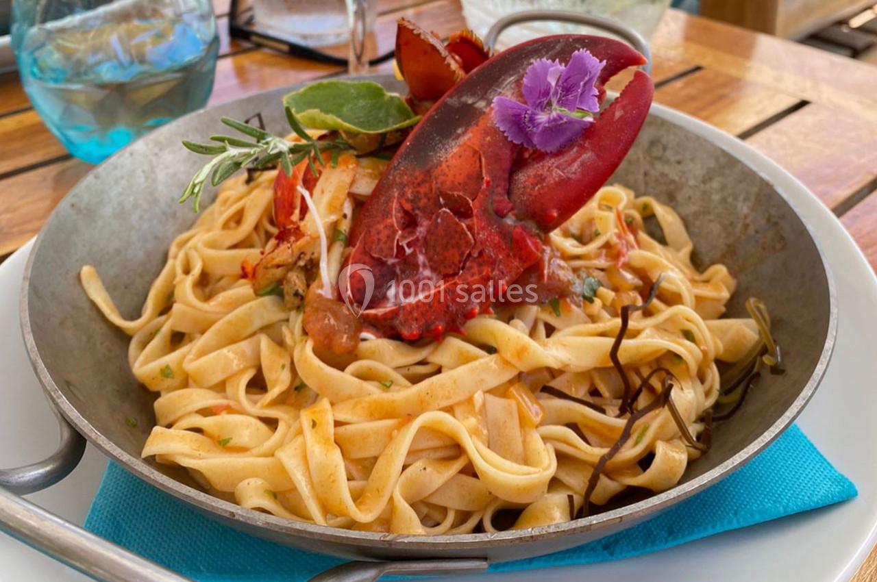 Assiette de tagliatelles garnie de morceaux de homard, herbes fraîches et fleur comestible, servie sur une table en bois.