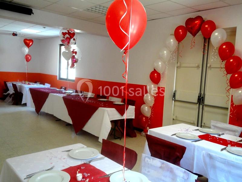 Salle décorée pour une fête avec des ballons rouges et blancs, nappes assorties et tables dressées.