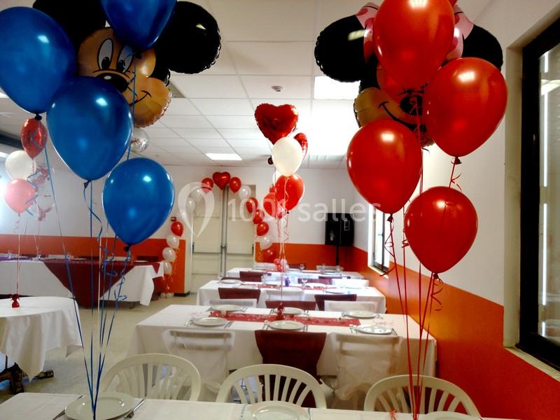 Salle décorée pour une fête avec des ballons rouges, bleus et en forme de Mickey, tables dressées avec nappes blanches.