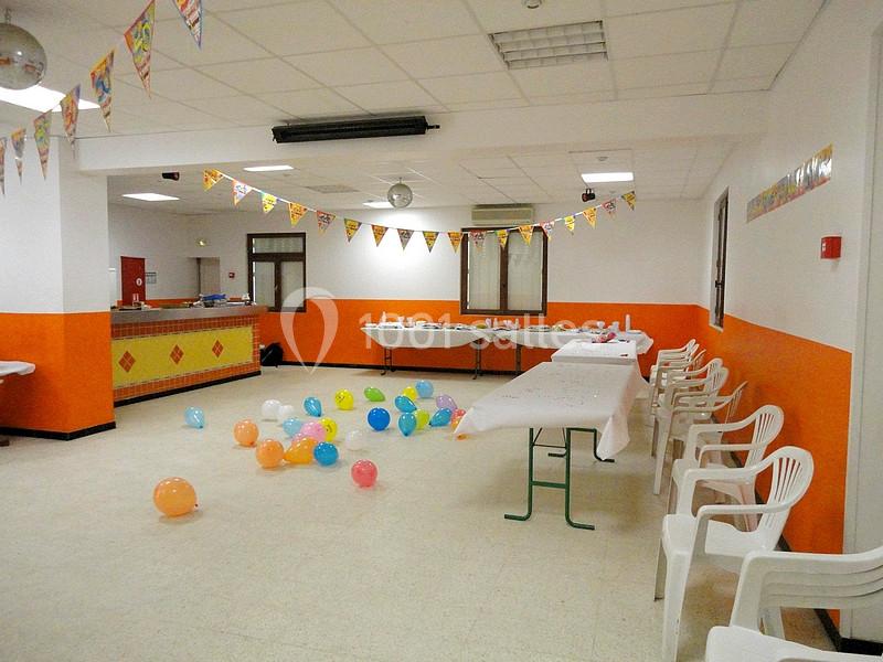 Salle décorée avec des guirlandes, des ballons colorés au sol, des tables dressées et des chaises en plastique alignées.