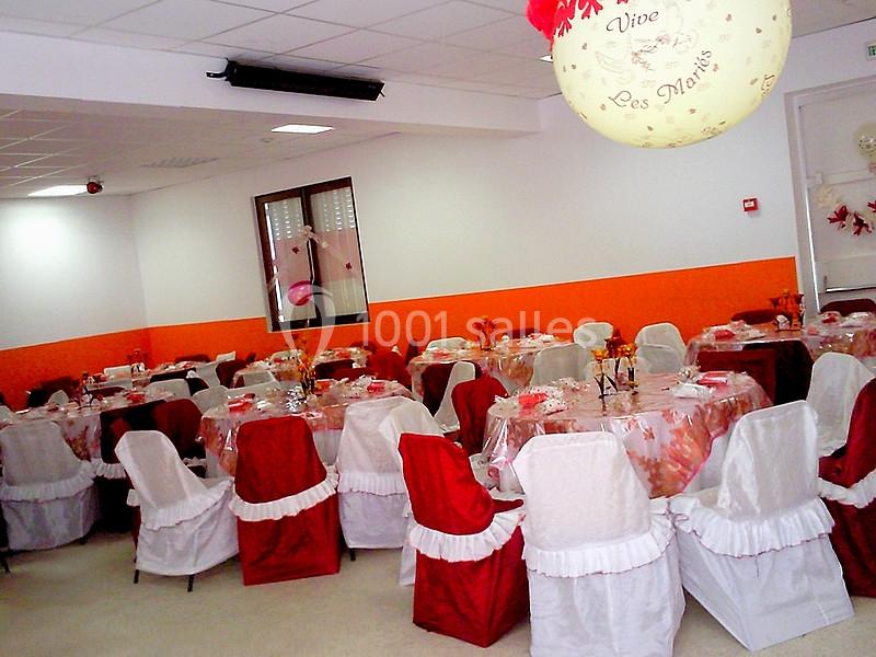 Salle décorée pour une réception, avec des tables dressées et des chaises recouvertes de housses rouges et blanches.