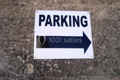 Panneau blanc avec le mot ’PARKING’ en noir et une flèche noire pointant vers la droite, fixé sur un sol en béton.