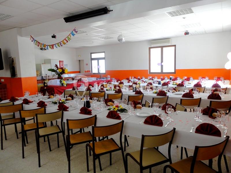Salle décorée pour une réception avec des tables dressées, nappes blanches, serviettes rouges et guirlande festive.