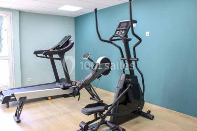 Deux équipements de fitness, un tapis de course et un vélo elliptique, dans une salle au sol en bois et murs bleus.