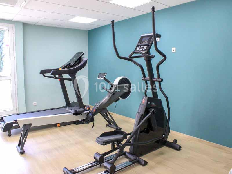 Deux équipements de fitness, un tapis de course et un vélo elliptique, dans une salle au sol en bois et murs bleus.