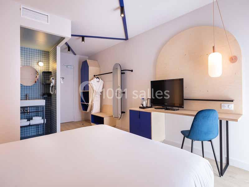 Chambre d'hôtel moderne avec lit, bureau, chaise bleue, télévision, salle de bain attenante et éclairage chaleureux.