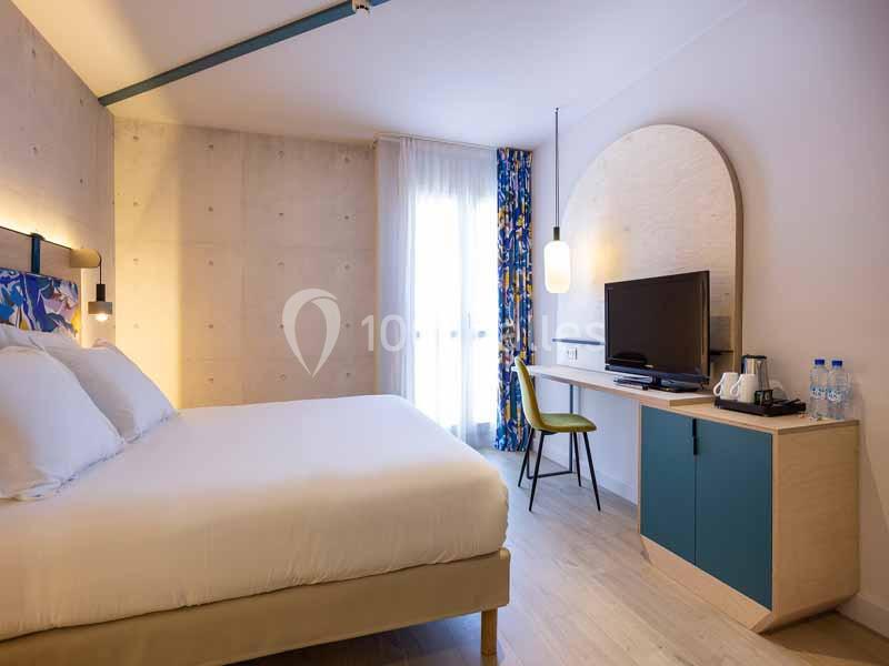 Chambre lumineuse avec lit double, bureau, télévision, bouilloire et fenêtre avec rideaux colorés.