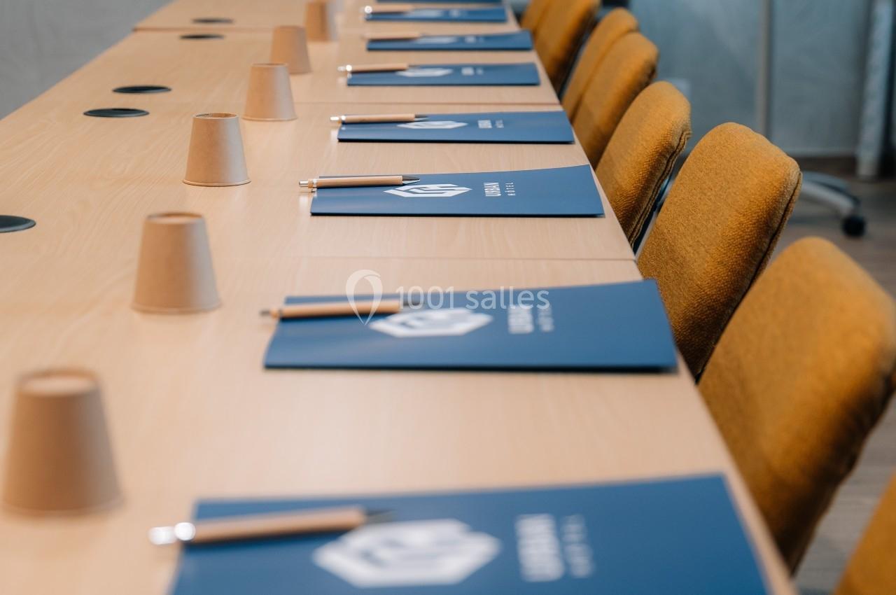 Salle de réunion avec une table équipée de carnets bleus, stylos et gobelets en carton alignés devant des chaises jaunes.
