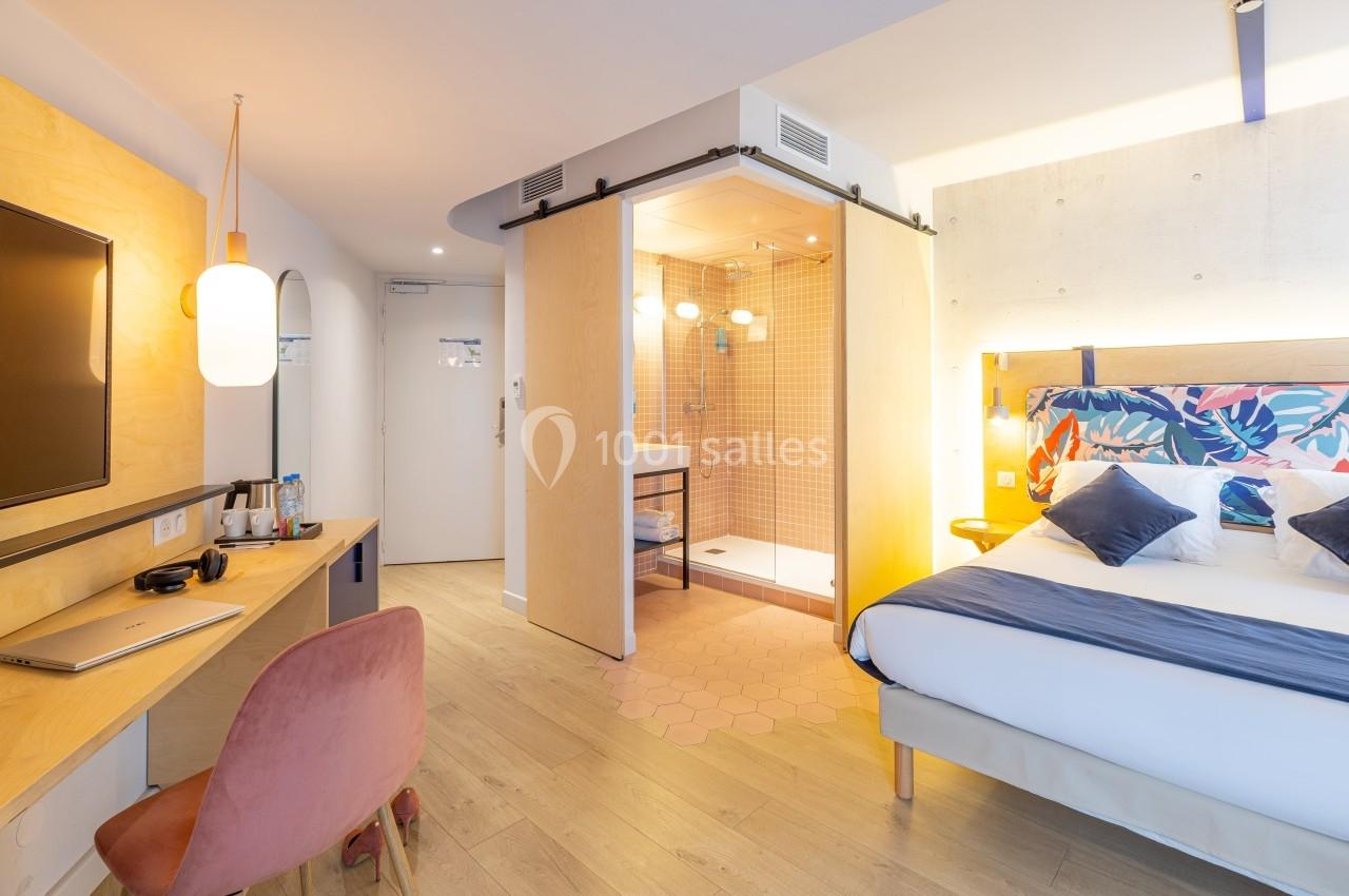 Chambre d'hôtel lumineuse avec lit double, bureau, télévision et salle de bain ouverte avec douche à l'italienne.