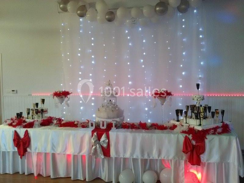 Table décorée pour une fête avec nappes blanches, rubans rouges, ballons, guirlandes lumineuses et une pièce montée centrale.