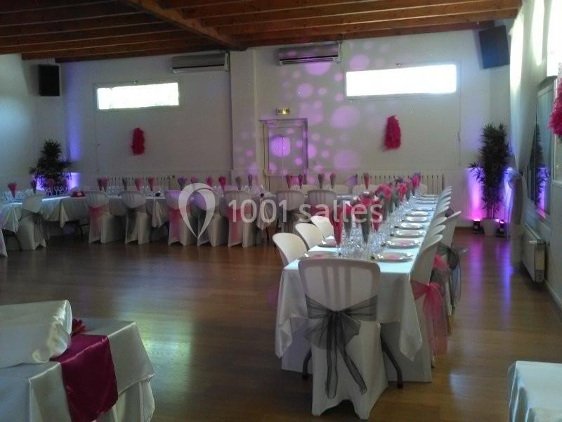 Salle de réception décorée avec des nappes blanches, des rubans roses et noirs, et des tables alignées pour un événement.