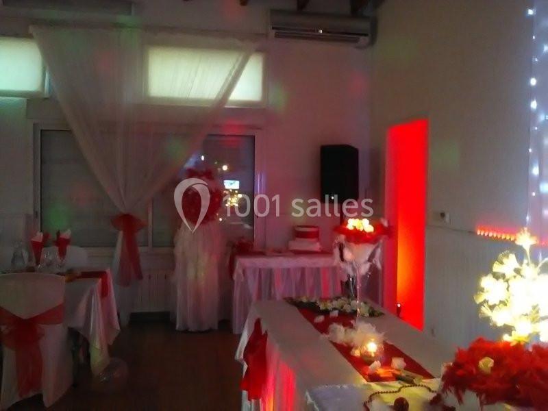 Salle décorée pour un événement, avec nappes blanches et rouges, éclairage tamisé et arrangements floraux.