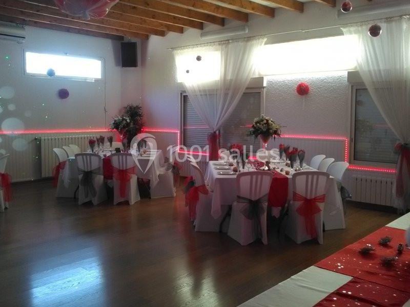Salle décorée pour un événement avec tables dressées, nappes blanches, chaises ornées de rubans rouges et éclairage tamisé.