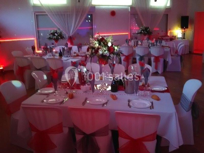 Salle de réception décorée avec des tables dressées, nappes blanches, chaises ornées de nœuds rouges et centre de table…