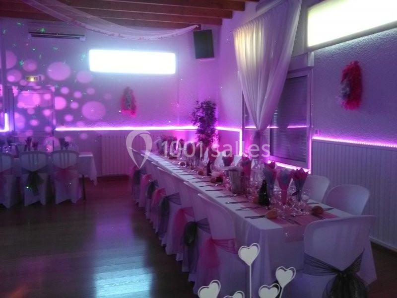 Salle décorée pour un événement, avec tables alignées, nappes blanches, chaises ornées de rubans et éclairage violet.