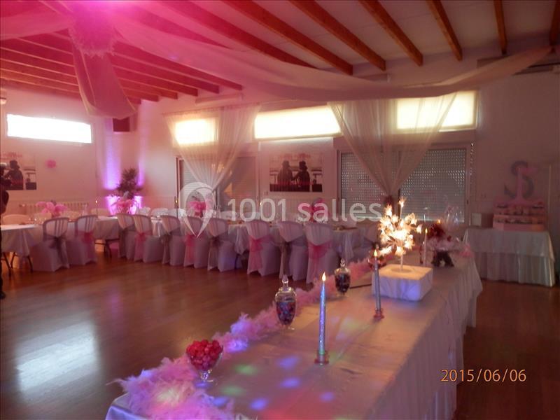 Salle décorée pour un événement festif avec tables dressées, nappes blanches, décorations roses et éclairage tamisé.