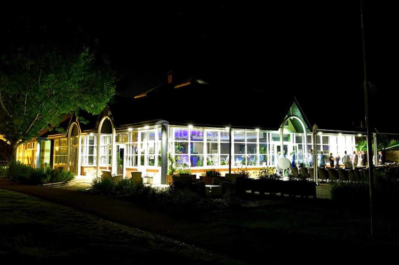 Bâtiment vitré éclairé de nuit, entouré d'arbres et de personnes visibles à l'intérieur.