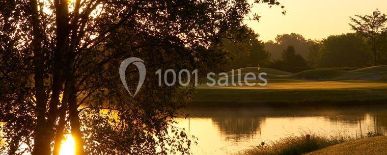 Paysage au lever du soleil avec un étang calme, des arbres en contre-jour et une pelouse vallonnée à l'arrière-plan.