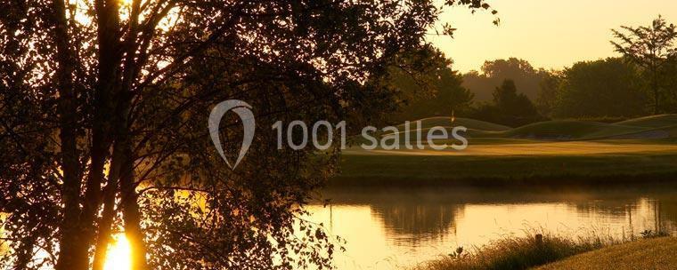 Paysage au lever du soleil avec un lac calme, des arbres en contre-jour et une pelouse vallonnée en arrière-plan.
