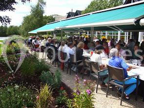 Terrasse d'un restaurant avec des clients attablés sous des stores verts, entourée de parterres fleuris.
