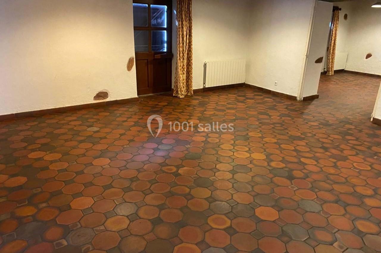 Salle vide avec sol en tomettes hexagonales rouges et brunes, murs blancs et porte en bois avec rideaux.