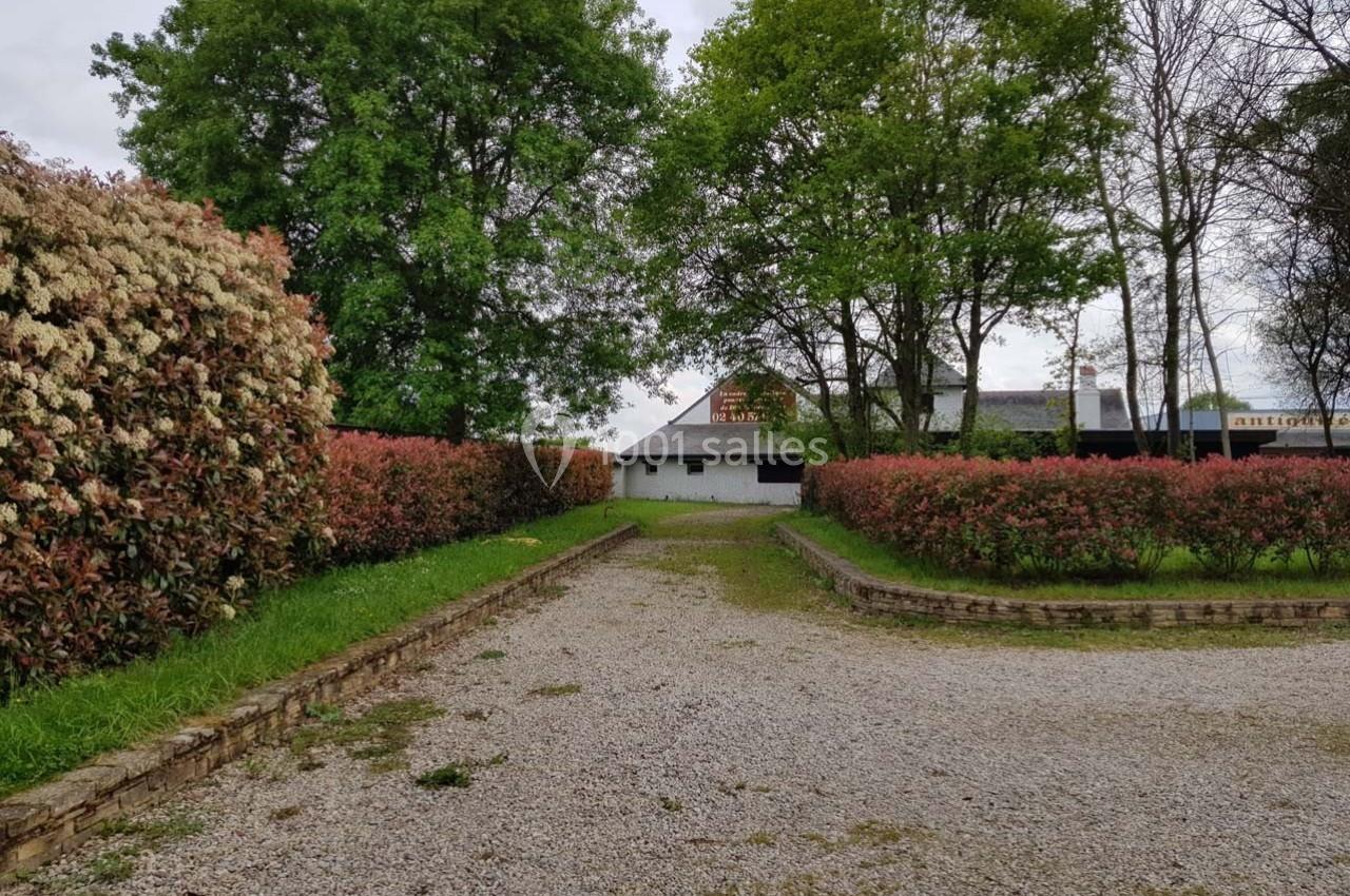 Allée en gravier bordée de haies fleuries menant à un bâtiment blanc entouré d'arbres.