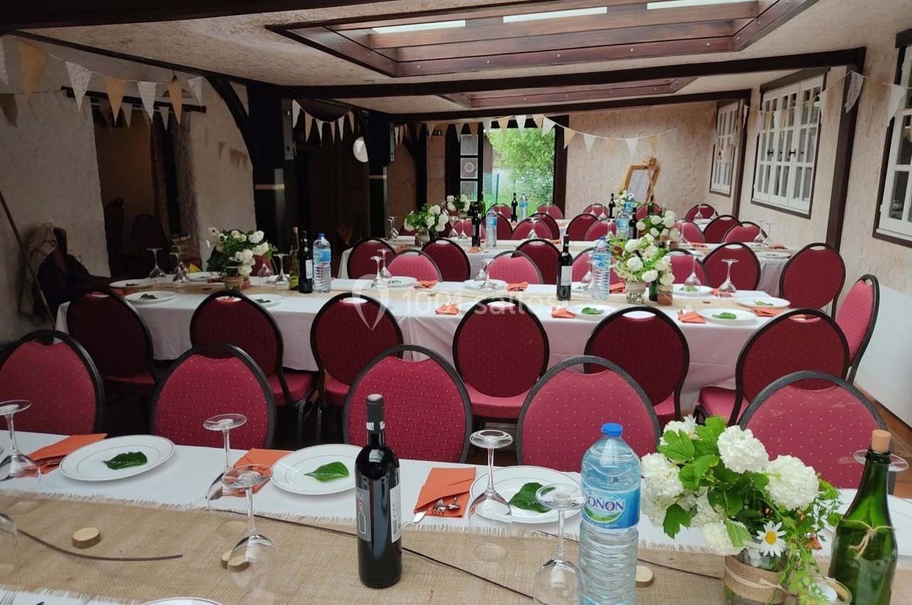 Salle décorée pour un repas festif avec tables dressées, chaises rouges, fleurs, bouteilles et guirlandes suspendues.