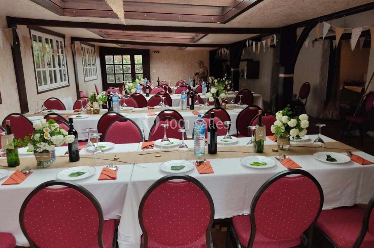 Salle décorée pour un événement avec des tables alignées, nappes blanches, chaises rouges et centres de table fleuris.