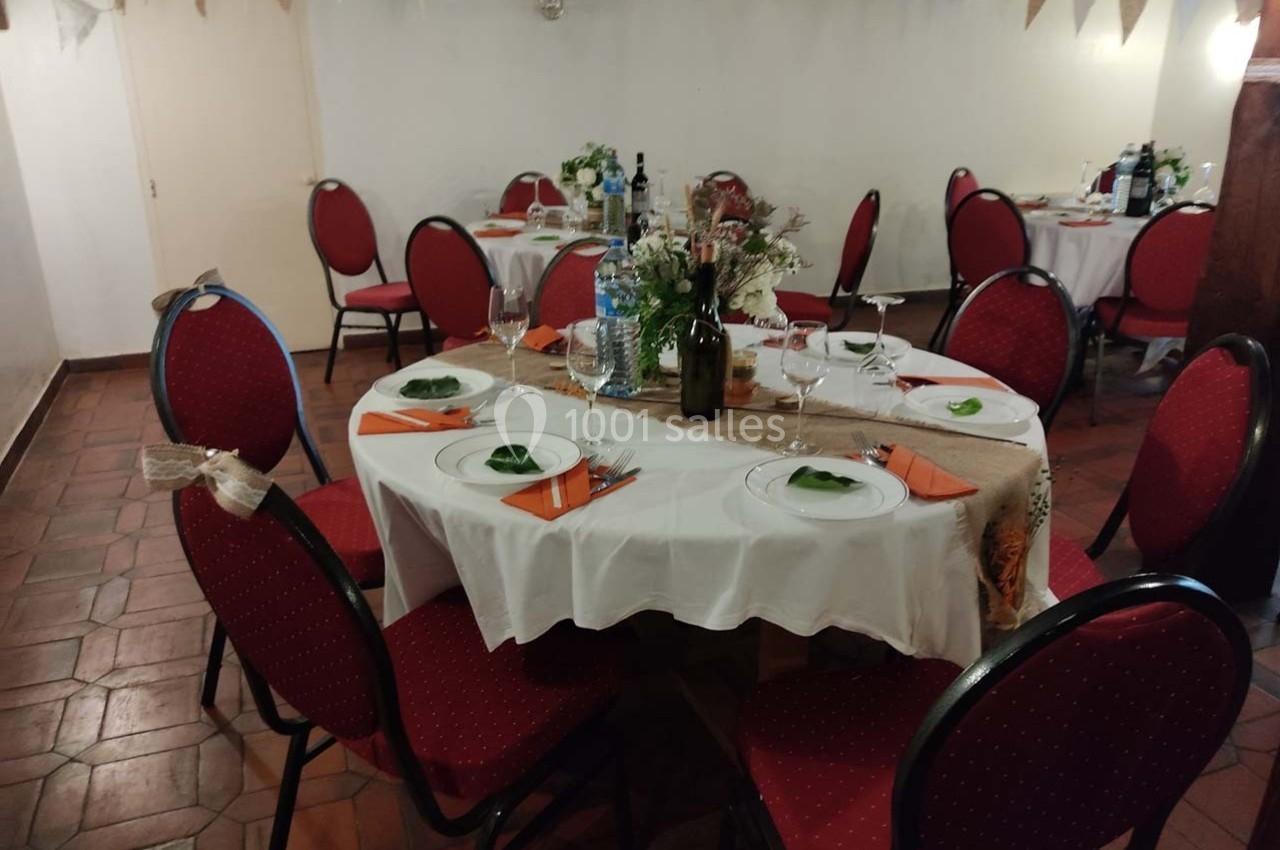 Salle décorée pour un repas, avec des tables rondes dressées, nappes blanches et chaises rouges.