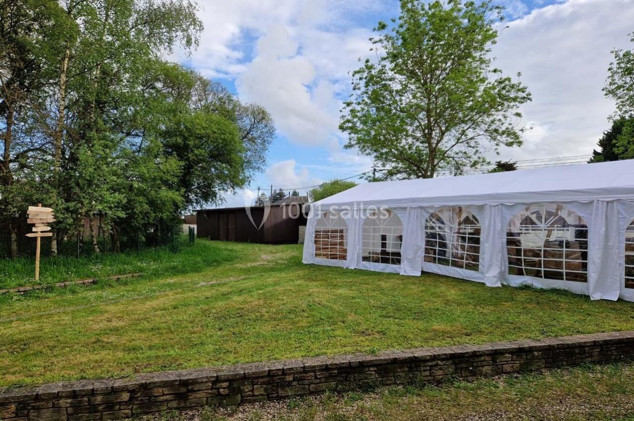 Tente blanche installée dans un jardin verdoyant avec des arbres et un ciel partiellement nuageux en arrière-plan.