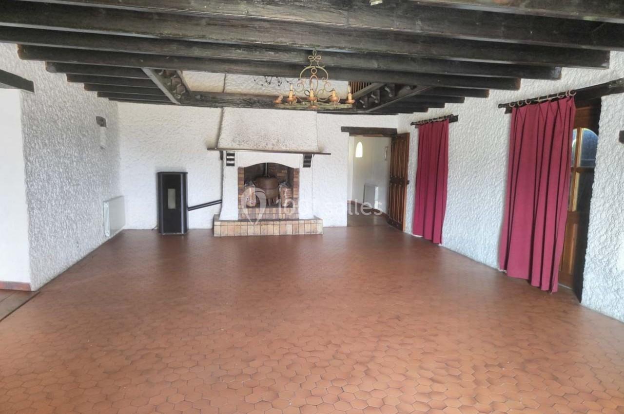 Grande pièce avec sol en carrelage hexagonal, murs blancs texturés, poutres en bois apparentes et cheminée en pierre.