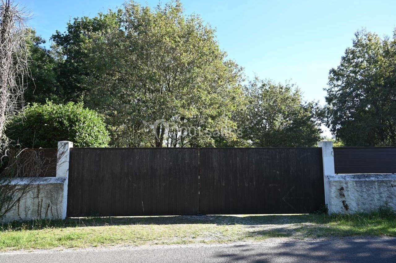 Portail en bois sombre fermé, encadré par des murs blancs, avec des arbres en arrière-plan sous un ciel dégagé.
