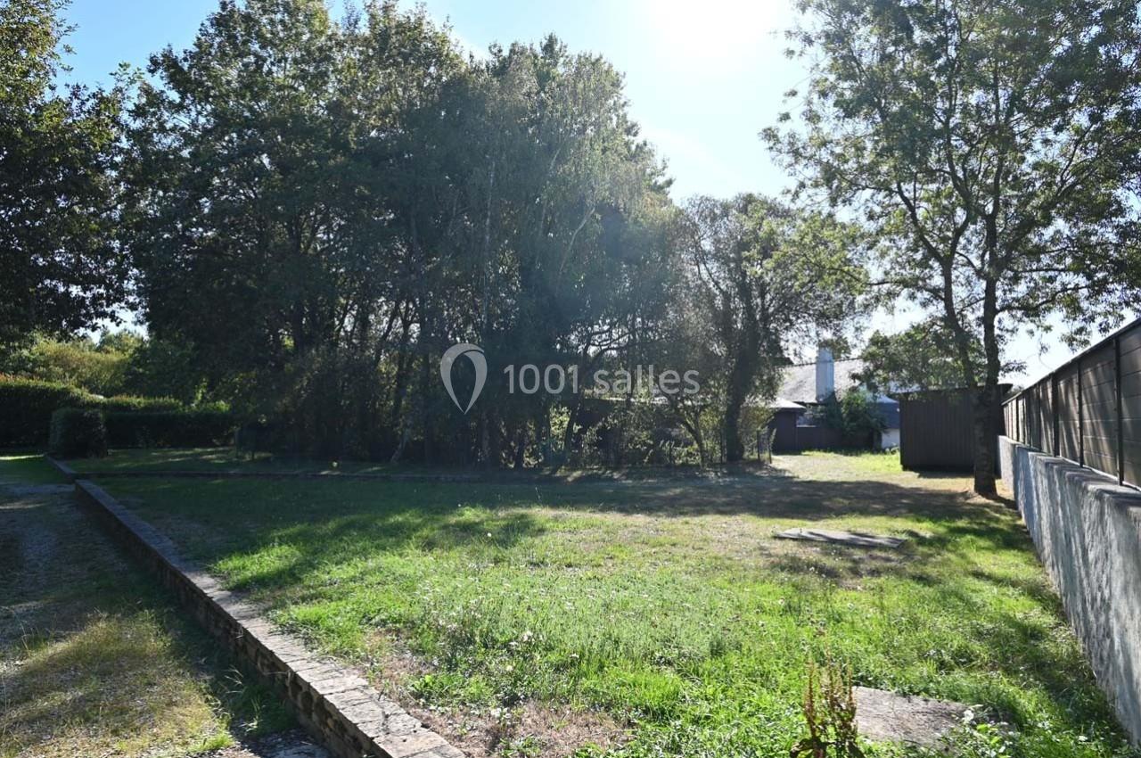 Terrain herbeux entouré d'arbres, avec des bâtiments en arrière-plan et une clôture sur la droite.