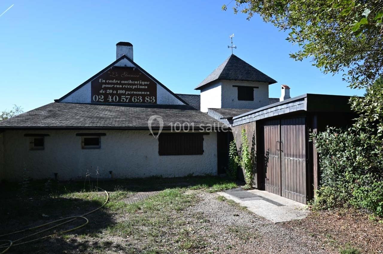 Bâtiment blanc avec toit en ardoise, garage en bois et enseigne publicitaire sur la façade, entouré de végétation.