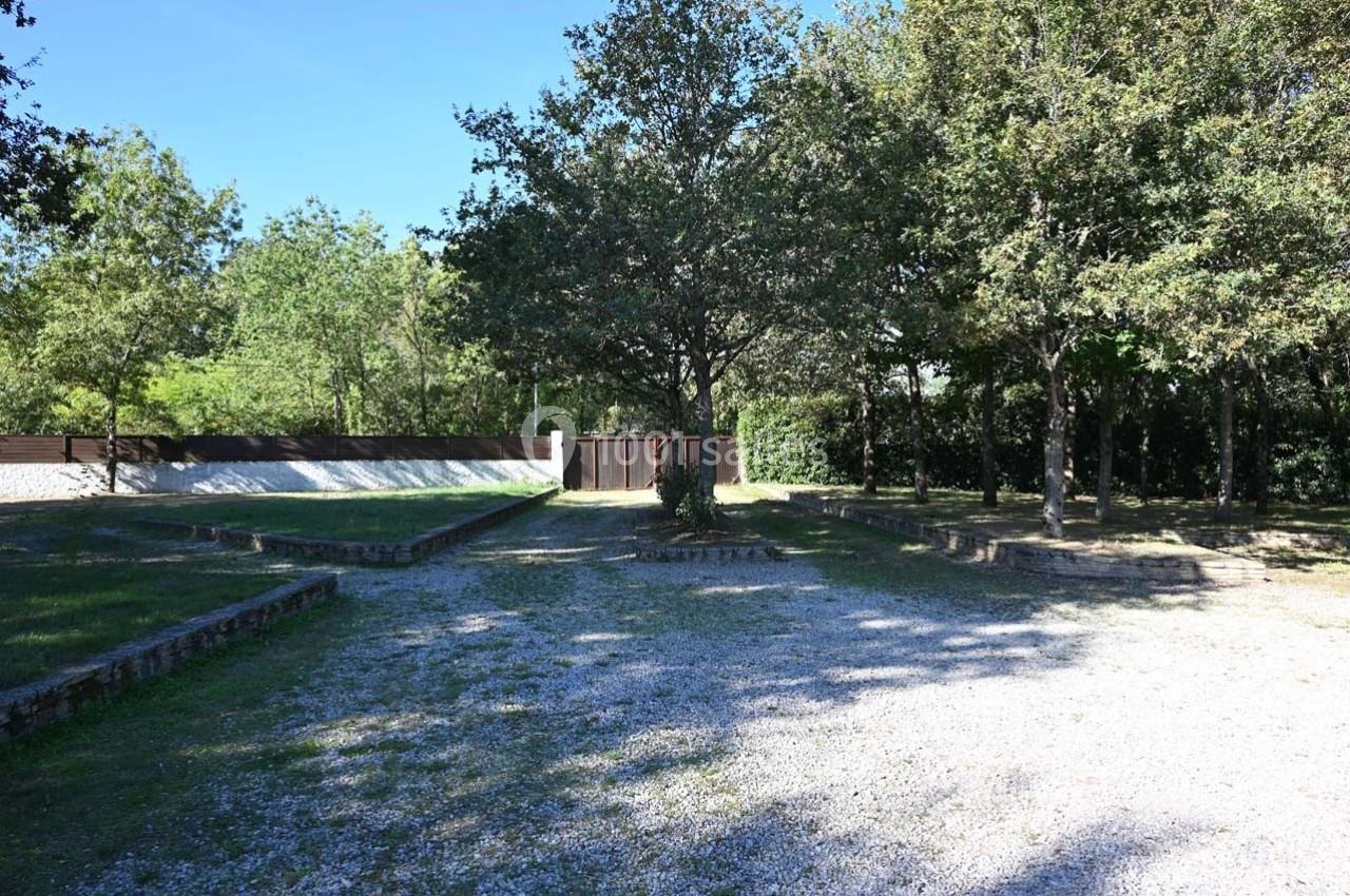 Allée gravillonnée bordée d'arbres et de murets en pierre, menant à un portail en bois fermé.