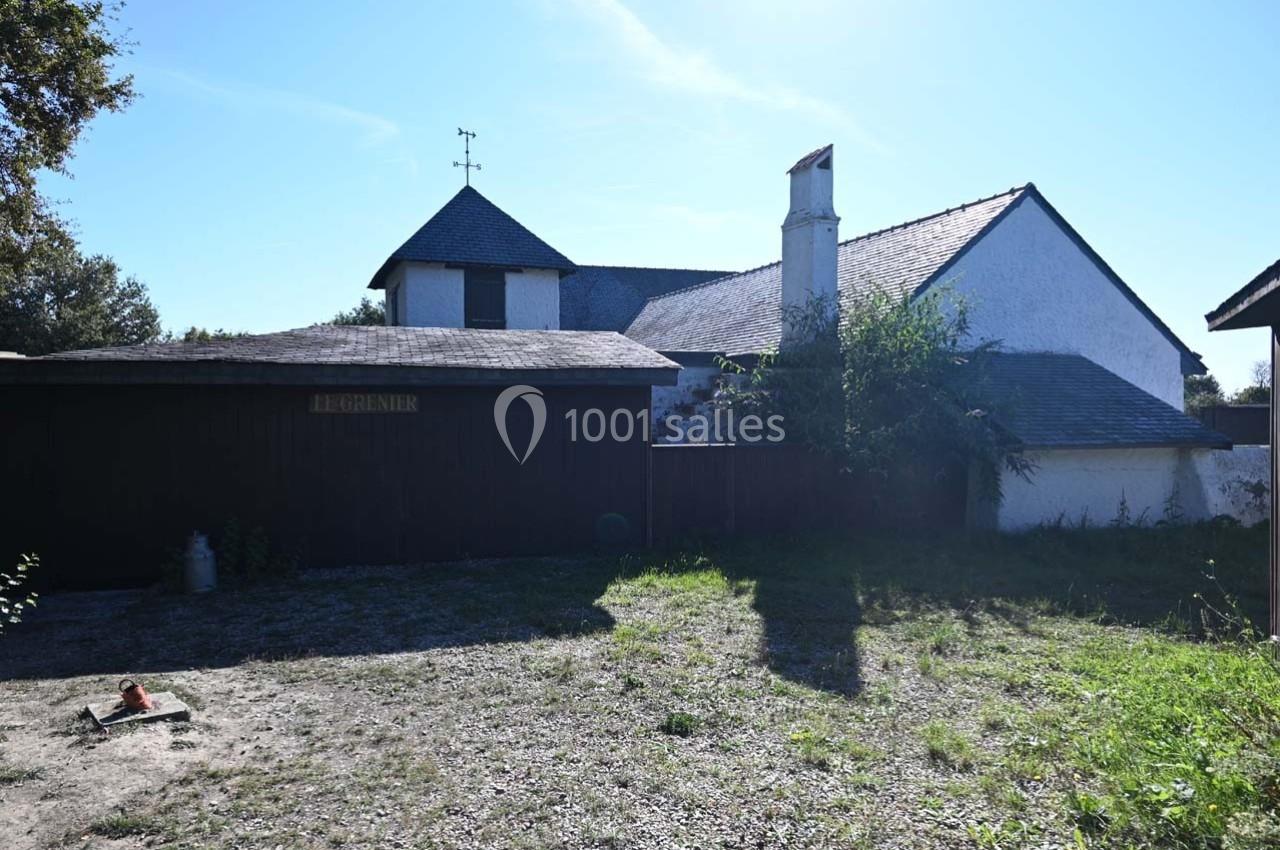 Cour extérieure d'une maison avec toit en ardoise, un bâtiment annexe en bois et un sol en gravier partiellement herbeux.