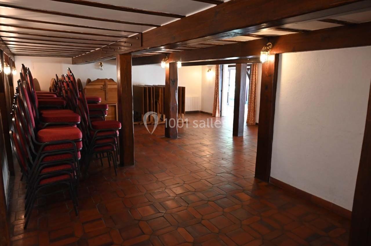 Salle vide avec sol en carrelage, poutres apparentes, chaises empilées et lumière naturelle entrant par une porte vitrée.