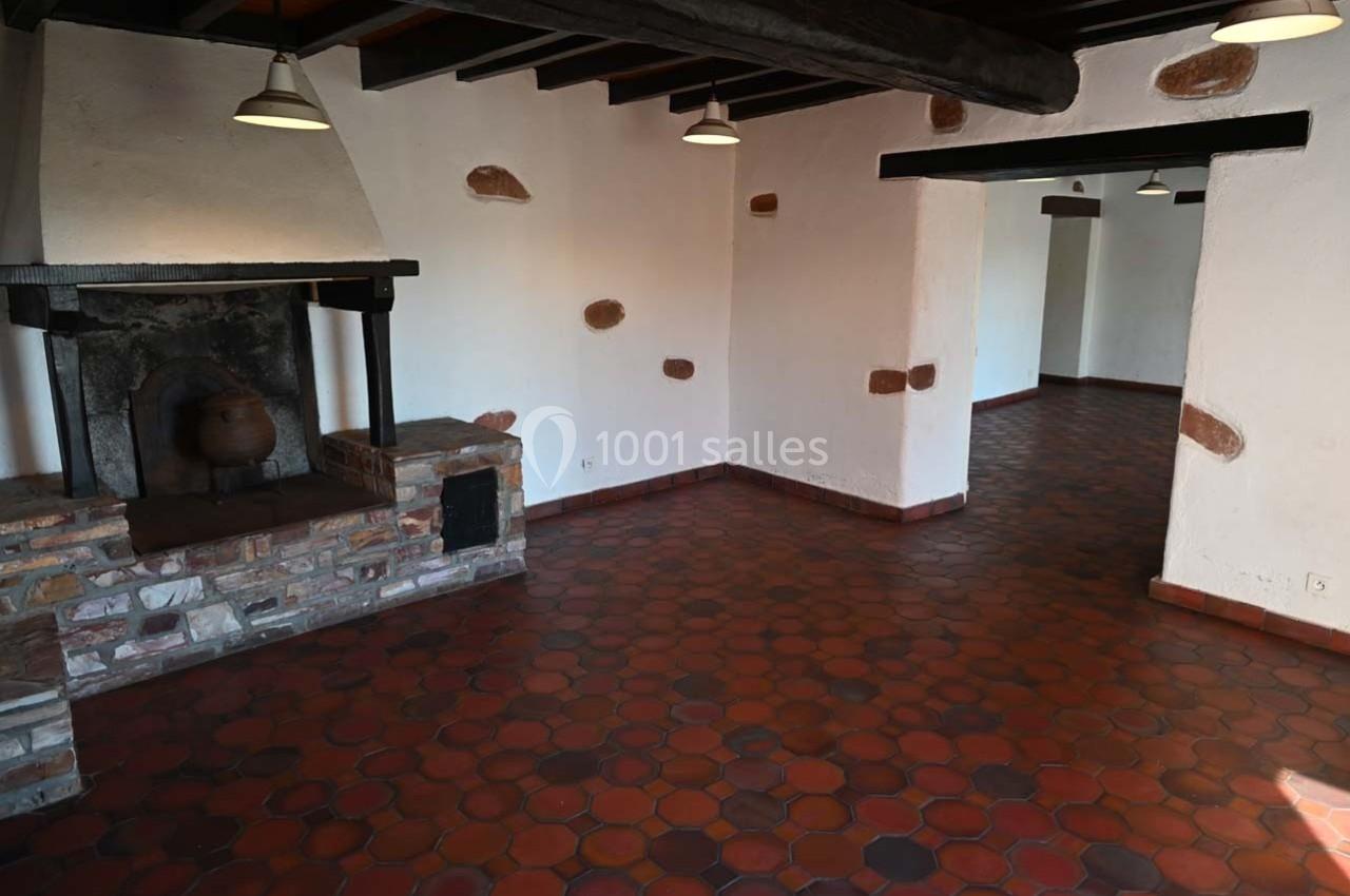 Intérieur d'une pièce vide avec sol en tomettes rouges, cheminée en pierre et poutres apparentes au plafond.