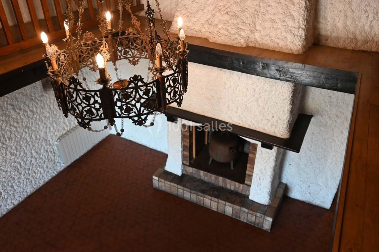 Vue intérieure d'une pièce avec cheminée en briques, murs texturés blancs, sol en carrelage rouge et lustre en métal.