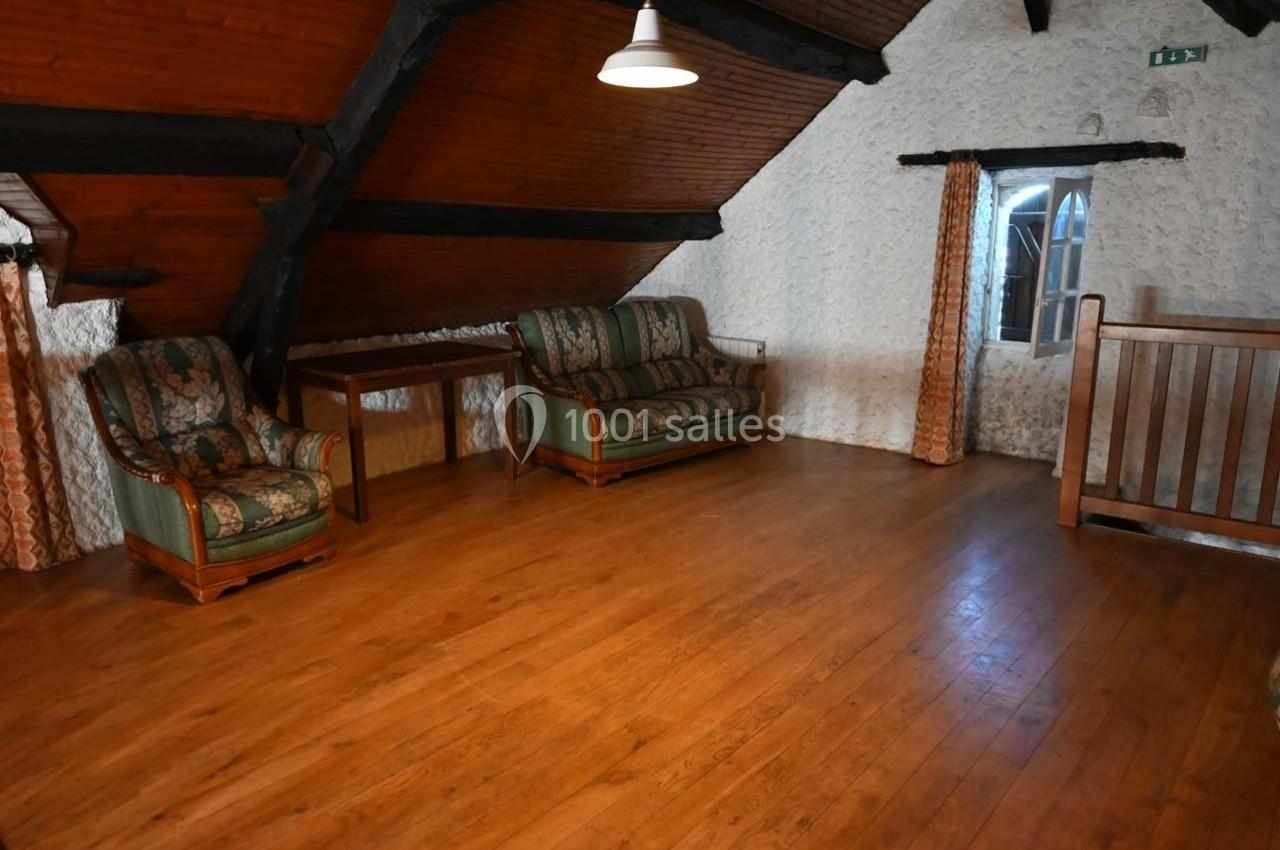 Salle mansardée avec parquet en bois, murs en pierre, mobilier ancien et une fenêtre avec rideaux.
