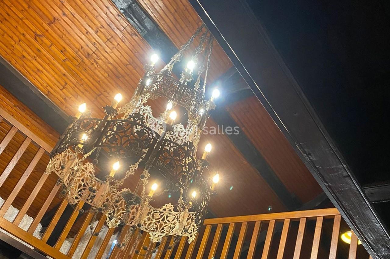 Lustre en métal orné suspendu à un plafond en bois, éclairé par plusieurs ampoules, dans un intérieur rustique.