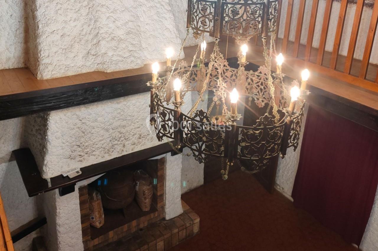 Lustre en métal forgé allumé, suspendu dans une pièce avec cheminée en pierre et mezzanine en bois.