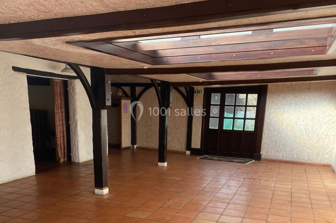 Salle vide avec sol en carrelage orange, poutres en bois sombre, murs clairs et porte vitrée donnant sur l'extérieur.