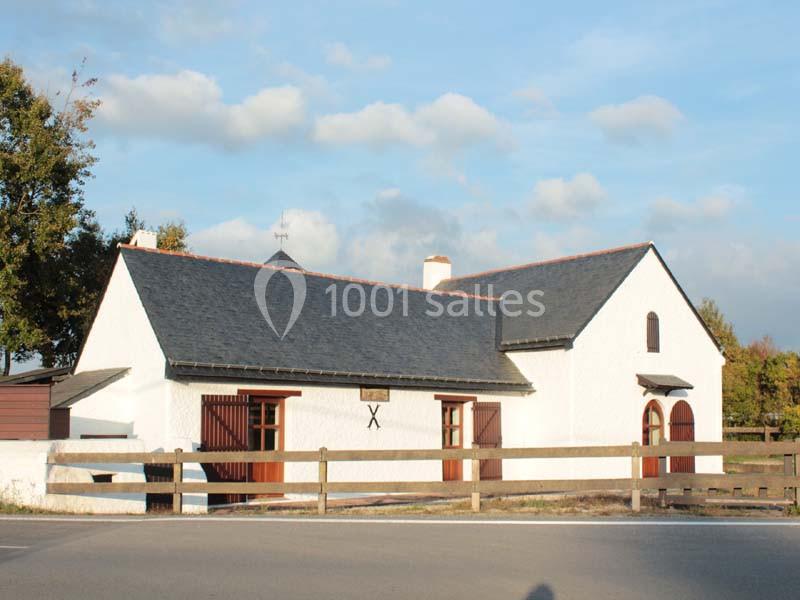 Bâtiment blanc à toit d'ardoise avec une clôture en bois, situé près d'une route sous un ciel partiellement nuageux.