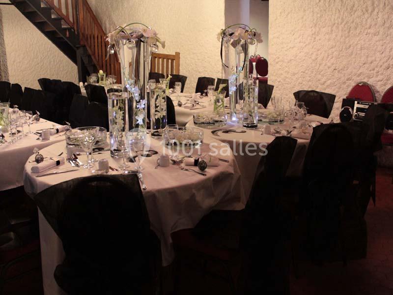 Salle décorée pour un événement avec des tables rondes dressées, nappes blanches et centres de table floraux.