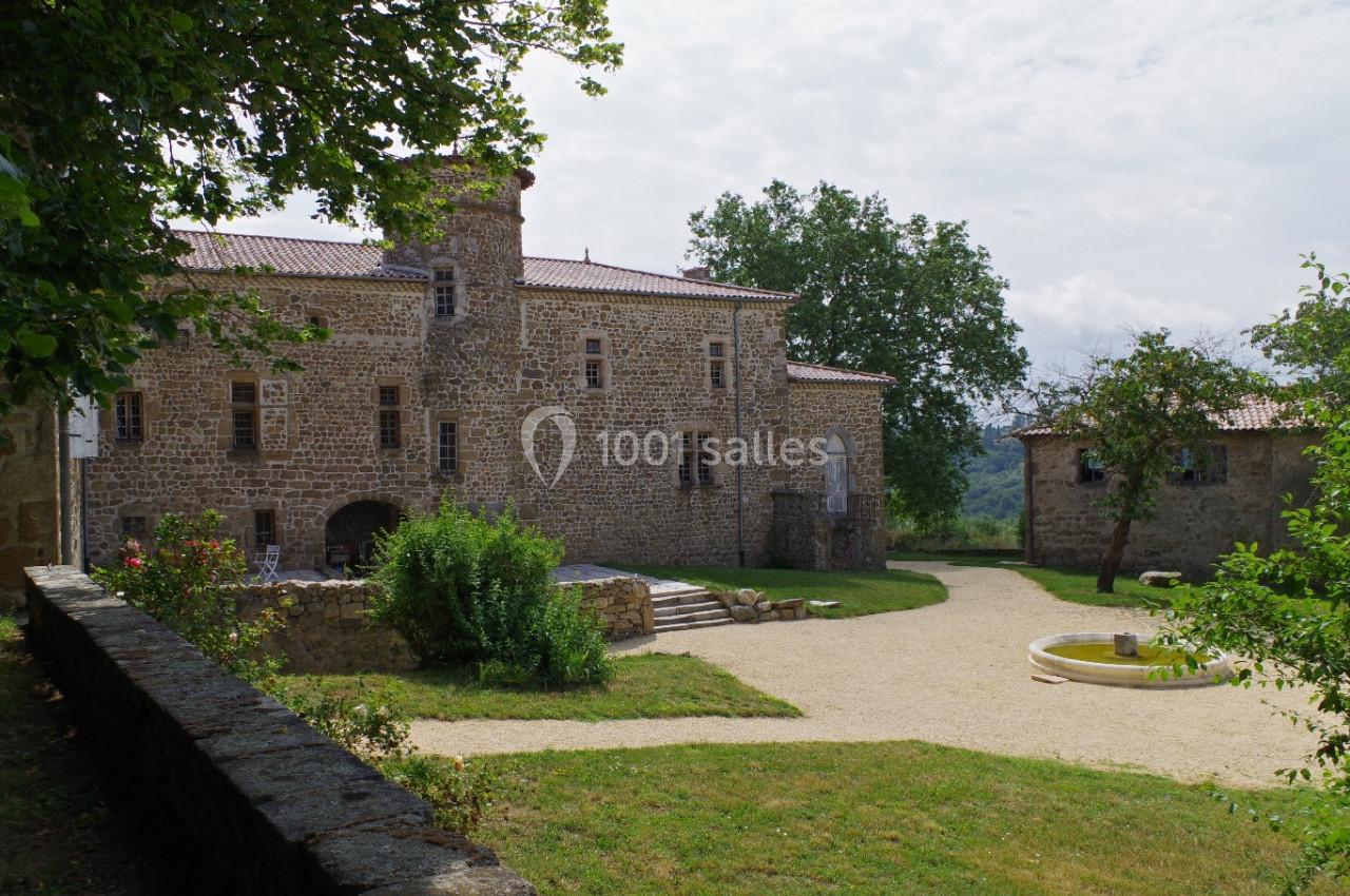 Cour d’un bâtiment en pierre avec une fontaine centrale, entourée de verdure et d’arbres sous un ciel partiellement nuageux.